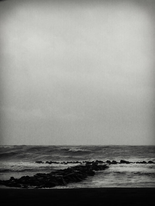 Scogliere artificiali avanzano in diagonale nel mare sotto un vasto cielo vuoto in bianco e nero. - Black-and-white seascape with artificial breakwaters extending diagonally beneath a vast, empty sky.