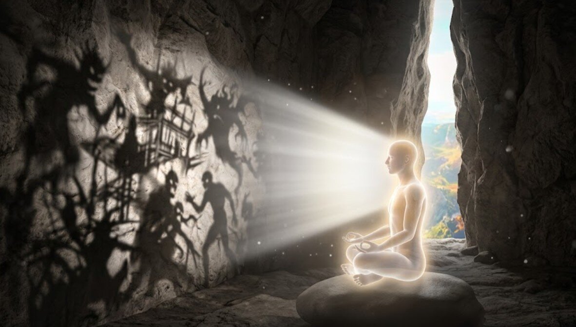 L'interno di una caverna di pietra oscura e grezza. Da una stretta fenditura rocciosa, la luce esterna proietta sulla parete opposta ombre confuse e mostruose che rappresentano i pensieri di paura. Al centro, una figura umana eterea e luminosa, seduta in meditazione profonda, osserva queste proiezioni con un sorriso sereno, non identificandosi con esse. La luce pura del proiettore divino brilla dietro di lei, rivelando l'illusione.