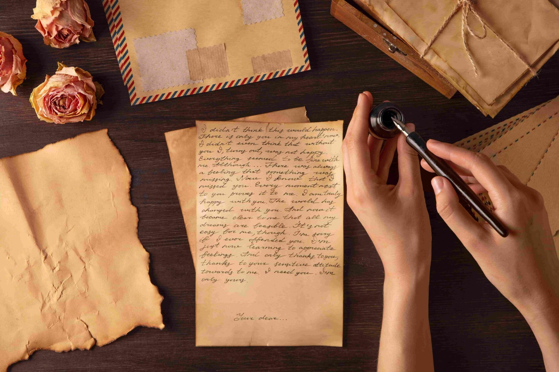 tavolo di legno con sopra carta da lettere, calamaio, rose secche e due mani che stanno per scrivere qualcosa di importante