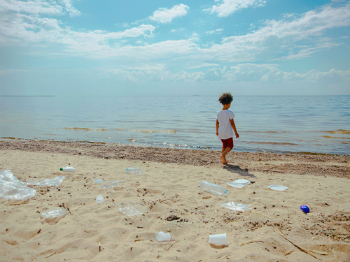 Bambino con capelli ricci in riva al mare con la spiaggia piena di bottiglie di plastica