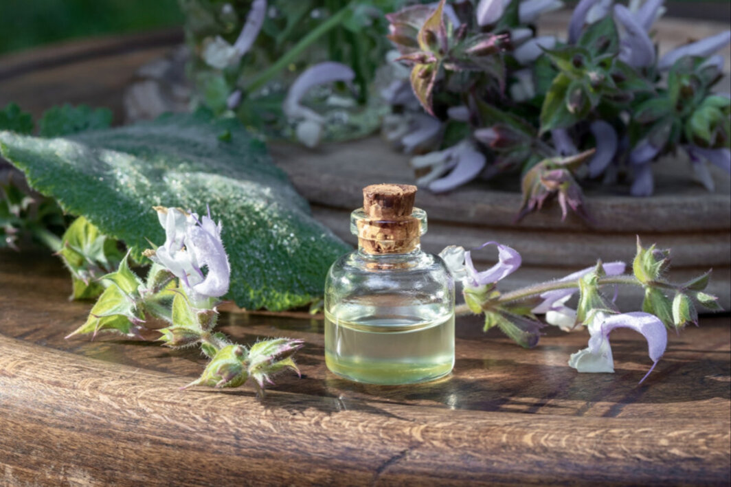Fotografia di una boccetta di vetro di olio essenziale con tappo in sughero, appoggiata su un tavolo di legno. Sullo sfondo bouquet di fiori e foglie di erbe officinali