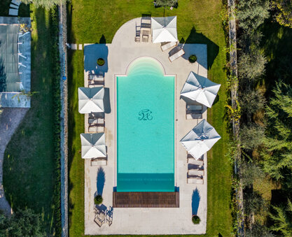 ripresa aerea zenitale di piscina rettangolare con lettini e ombrelloni bianchi circondata dal prato