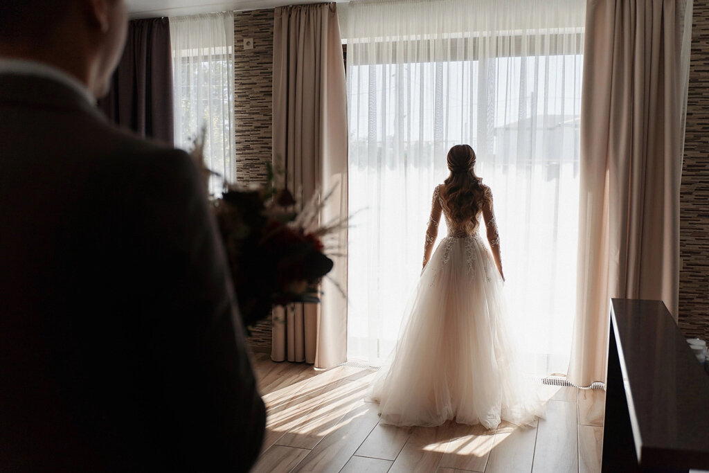 sposa in abito bianco in controluce che guarda fuori dalla finestra mentre lo sposo la osserva in secondo piano
