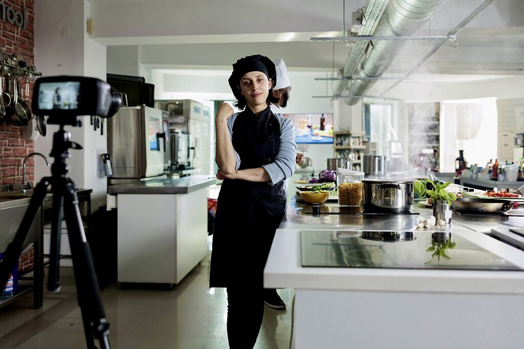 chef che posa in una cucina professionale mentre viene ripreso da una videocamera su treppiede