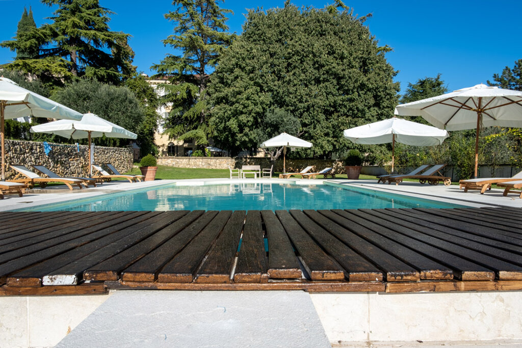 piscina esterna con bordo in legno, lettini e ombrelloni bianchi in una struttura immersa nel verde
