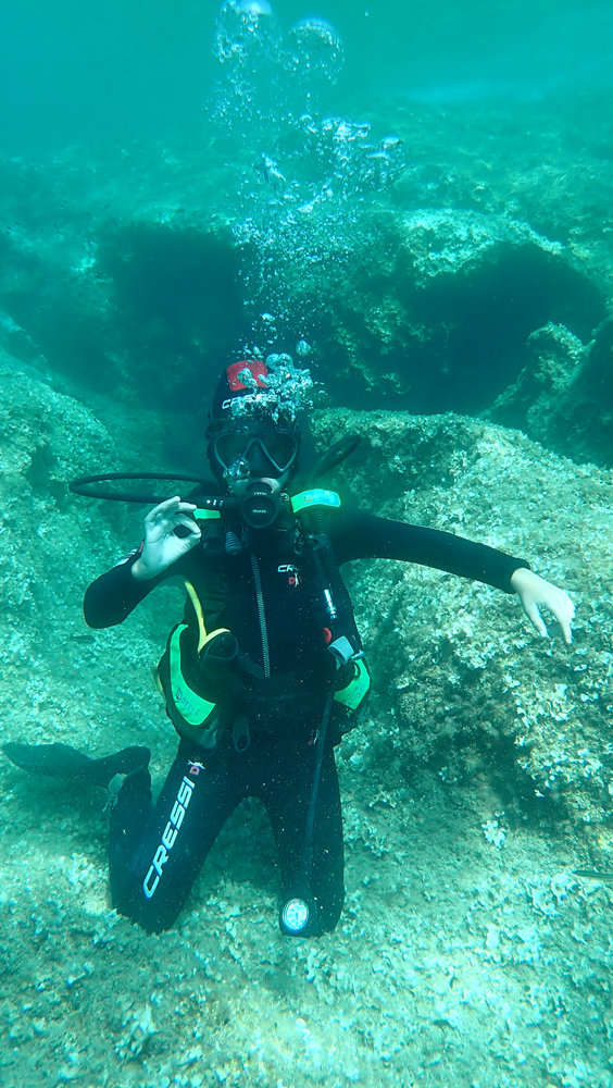immersione-sub-acqua-smeraldo-sardegna.png Il giovane Hugo saluta felice la fotocamera durante una splendida immersione subacquea nelle acque verde smeraldo della Sardegna.
