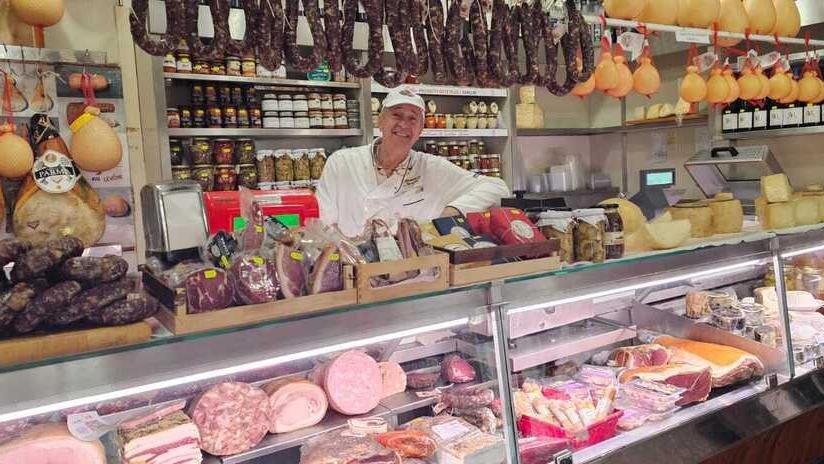Pinuccio Mangatia in primo piano nel suo storico negozio a Sassari, davanti a una ricca esposizione di salumi artigianali e formaggi sardi.