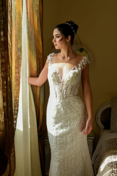 Bellissima sposa guarda dolcemente verso la finestra mentre un magico fascio di luce illumina parzialmente il suo vestito.