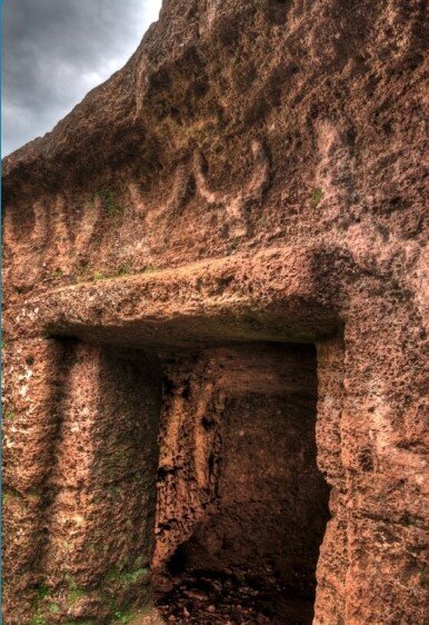 necropoli-anghelu-ruju-alghero-visita.jpeg Fotografia del portale d’ingresso scavato nella roccia arenaria di una domus de janas nella necropoli di Anghelu Ruju ad Alghero. L’immagine mostra l’architrave monolitico decorato con sculture simboliche di corna taurine prenuragiche.