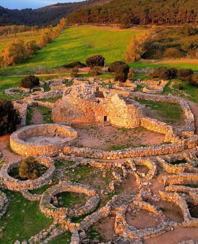 nuraghe-palmavera-alghero-sardegna.jpeg Fotografia panoramica aerea dall’alto del complesso nuragico Palmavera di Alghero. La vista mostra la struttura completa con il mastio centrale, le torri secondarie, l’antemurale e le fondamenta delle capanne del villaggio circostante immerse nella macchia mediterranea.
