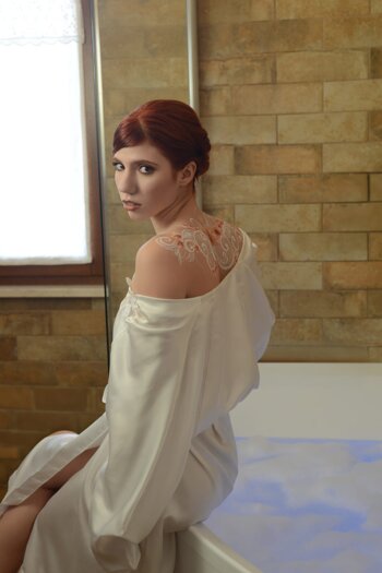 Sposa in vestaglia di seta sul bordo della vasca idromassaggio celeste con un calice di spumante Akenta e una candela rilassante.