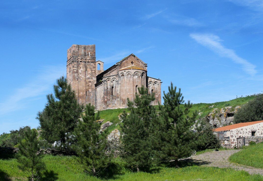 basilica-sant-antioco-di-bisarcio-sardegna-ozieri.jpeg Fotografia diurna della Basilica di Sant’Antioco di Bisarcio che svetta su una verde collina vulcanica nel territorio di Ozieri. L’immagine cattura la maestosa struttura medievale in pietra vulcanica isolata nel paesaggio rurale sardo.
