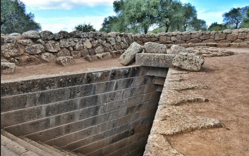 ingresso-pozzo-sacro-santa-cristina-sardegna.jpeg Fotografia reale diurna dell’ingresso del Pozzo Sacro di Santa Cristina a Paulilatino, Sardegna. L’inquadratura dall’alto mette in evidenza la straordinaria precisione architettonica nuragica e la simmetria della scala monumentale in basalto che scende verso la camera sotterranea.