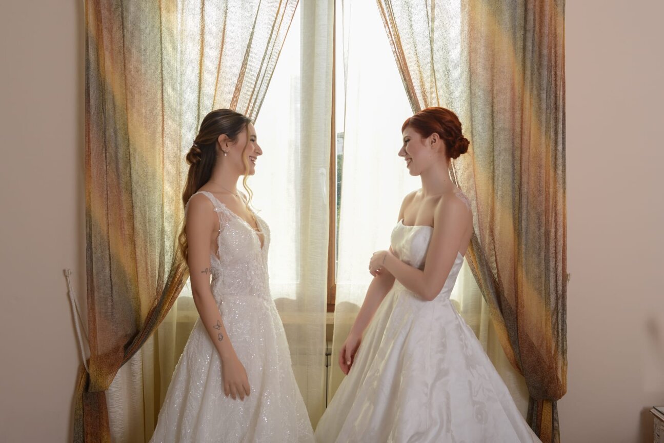 Due modelle in elegante abito da sposa si guardano complici nella luminosa camera matrimoniale di Villa Malvasio con la finestra sullo sfondo. Due modelle in elegante abito da sposa si guardano complici nella luminosa camera matrimoniale di Villa Malvasio con la finestra sullo sfondo.
