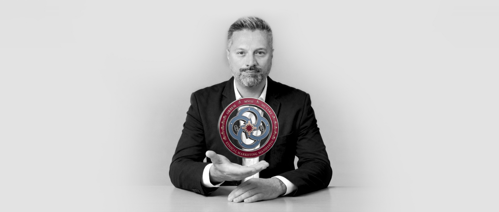  Barberis Federico Ottavio, seduto alla scrivania con una mano rivolta in camera che sostiene in sospensione il simbolo del Manifesto del Marketing Etico da lui creato.