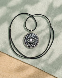 collana con pendente circolare, montatura argentata e decorazione mandala nei toni del bianco, nero e verde, sempre con cordino nero. Anche qui fondo verde salvia e luce naturale.