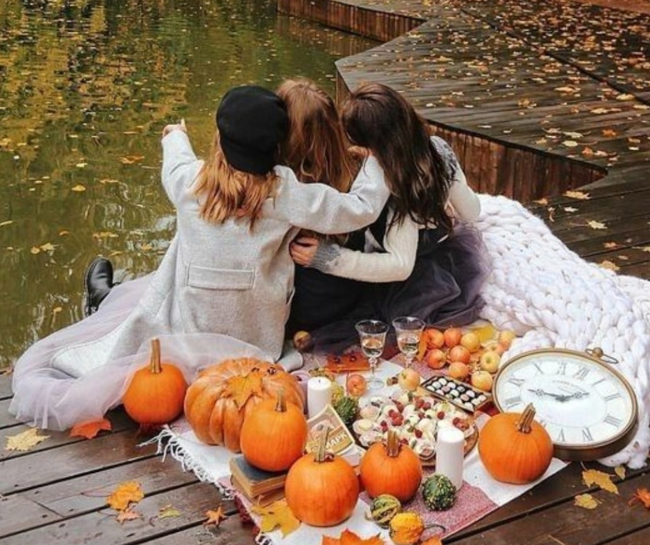 Tre donne abbracciate durante un picnic autunnale accanto a un lago, simbolo delle relazioni autentiche e del calore in stile Hygge