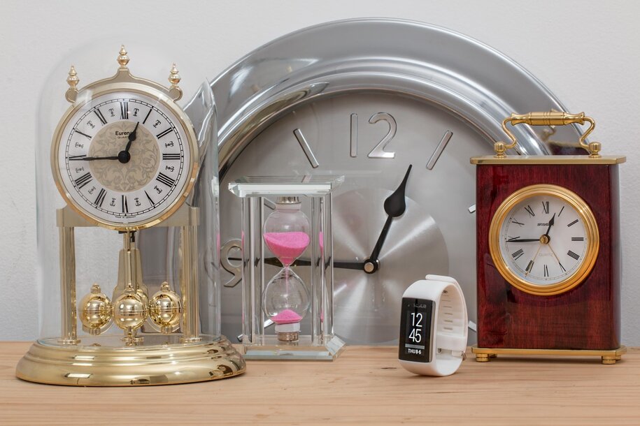 orologio a clessidra rosa, in primo piano orologio da polso bianco e orologio a campana e sullo sfondo orologio rotondo in acciaio e orologio da tavolo in legno su base di legno